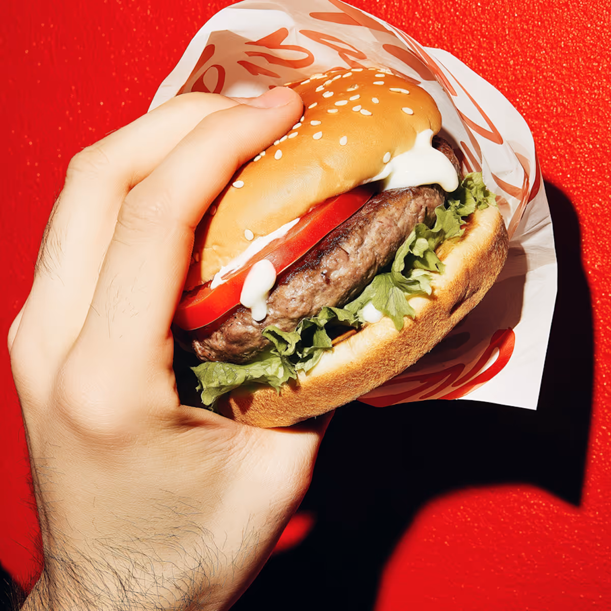 Hand holding a hamburger with lettuce, tomato, beef patty, mayonnaise, and sesame seed bun wrapped in paper against a red background.