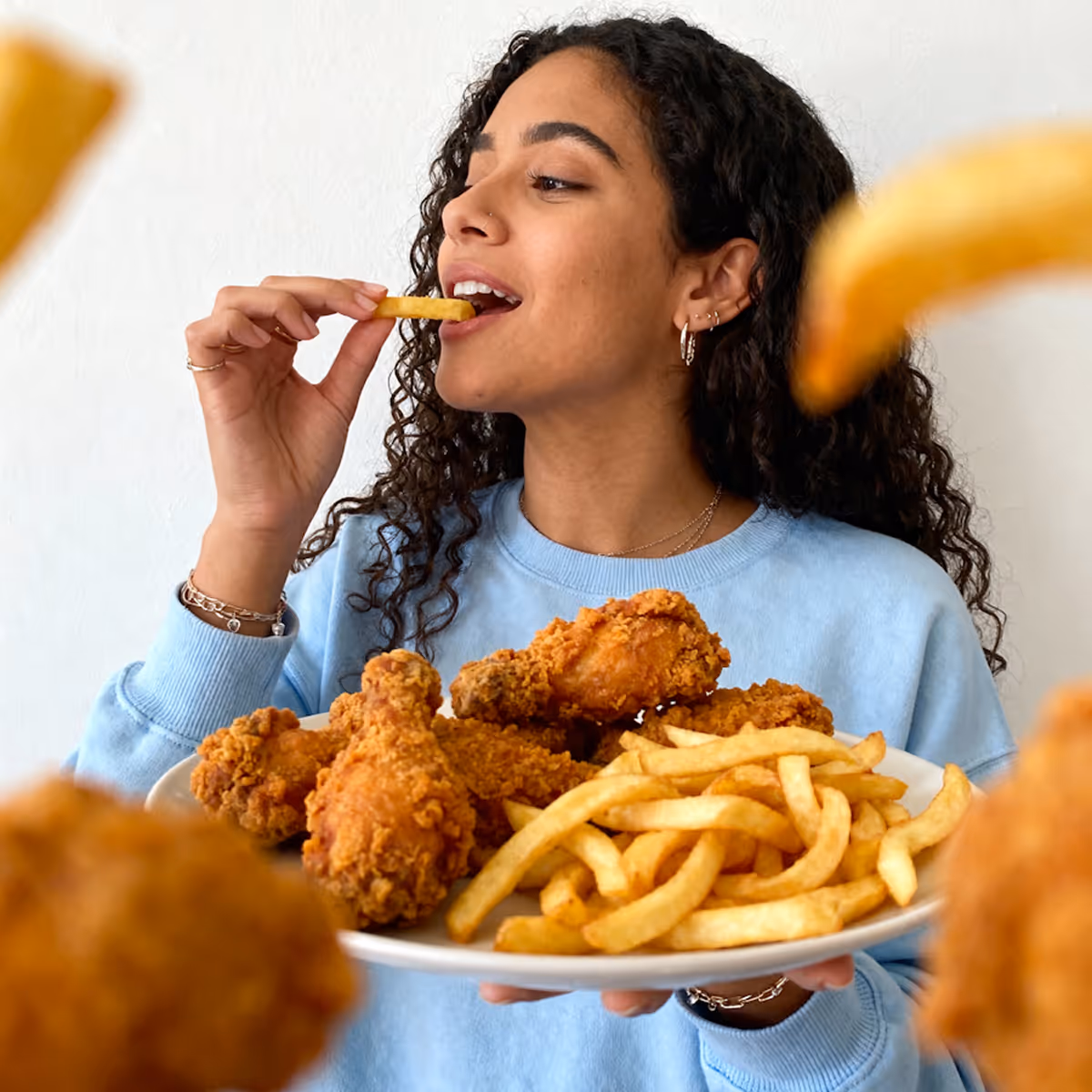 Woman in a blue sweater eating a french fry while holding a plate of fried chicken and french fries.